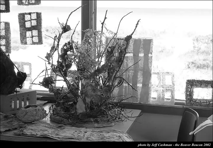 Black-and-white photograph of a branch-like paper sculpture displayed on a table beside a bright window.