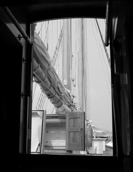 Black-and-white view through a vessel opening toward a mast, rigging, and wooden deck structures.