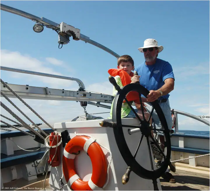 A child and an adult stand at the large steering wheel on a sailboat deck.