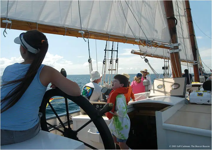 Children and adults gather on a sailing vessel’s deck, with one child at the large steering wheel.