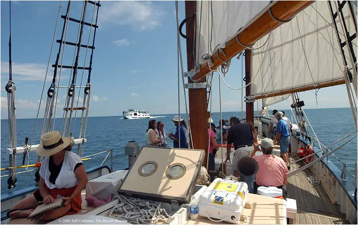 People gather on the deck of a sailing vessel beneath its rigging, with another boat visible offshore.