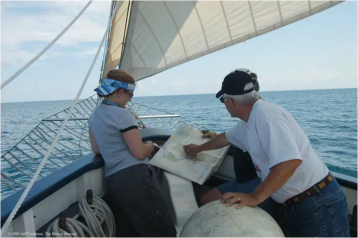 Two people consult a nautical chart aboard a sailboat under raised sails.