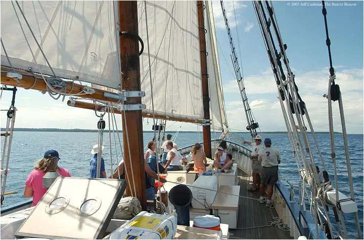 Passengers gather on the deck of a sailing vessel beneath partially raised sails.