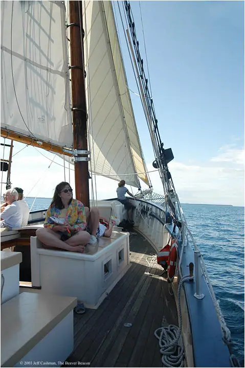 Passengers sit on the deck of a sailing vessel beneath billowing white sails.
