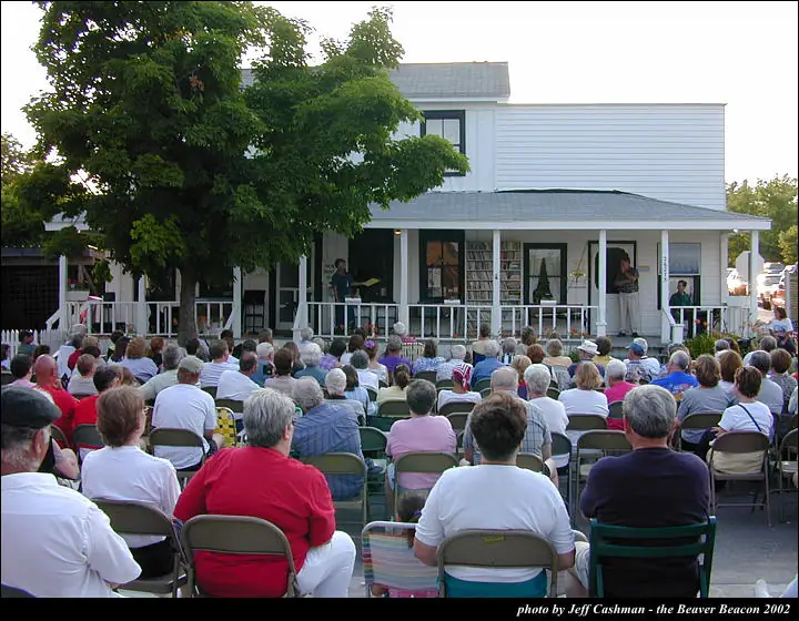 A large seated crowd watches people gathered on the porch of a white two-story building.