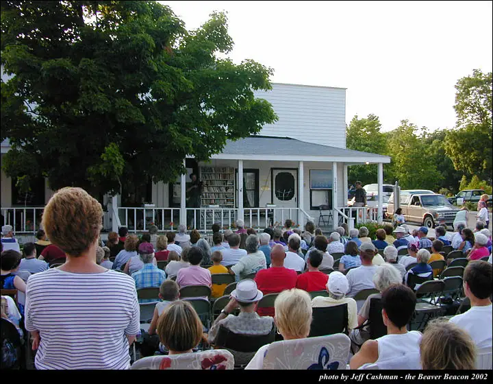 A large crowd sits in folding chairs facing people gathered on the porch of a small white building.