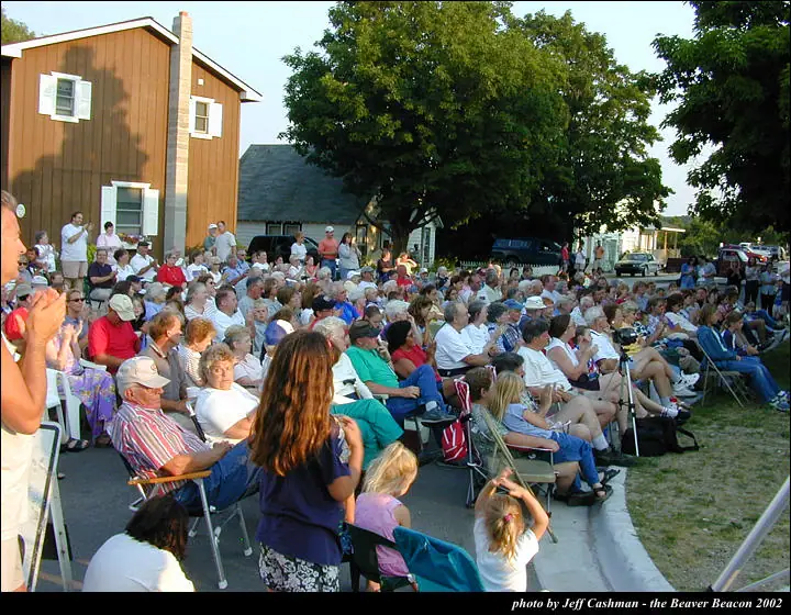 A large outdoor audience sits in folding chairs beside wooden buildings and trees.