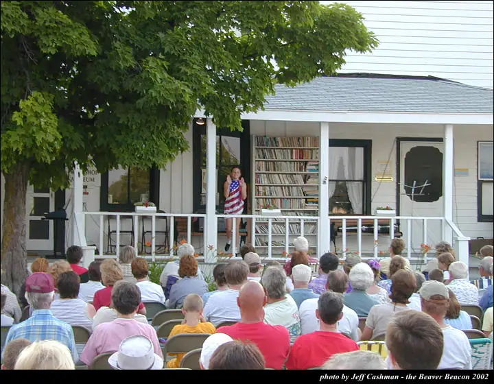 A woman in a flag-patterned dress addresses a large seated audience from a porch lined with bookshelves.