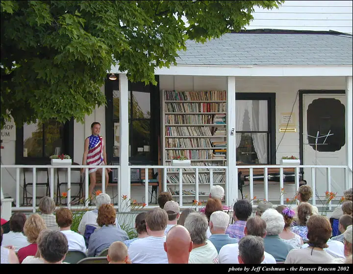 A woman in a flag-patterned dress stands on a porch before a seated audience.