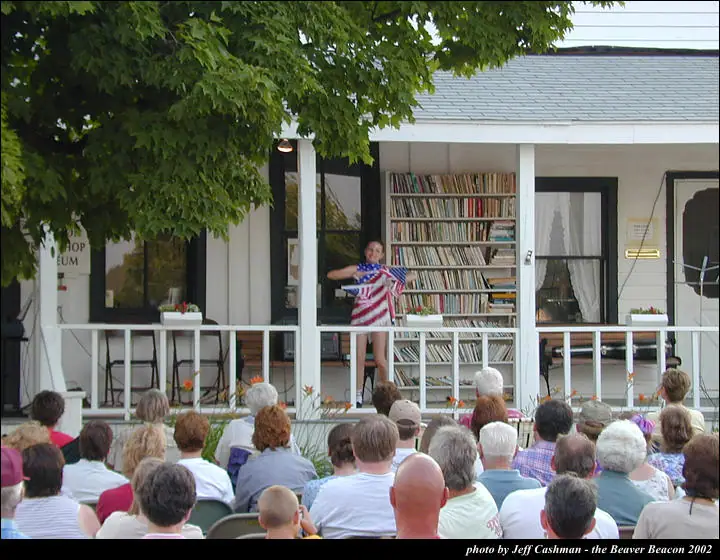 A woman in flag-patterned clothing performs on a porch before a seated audience.