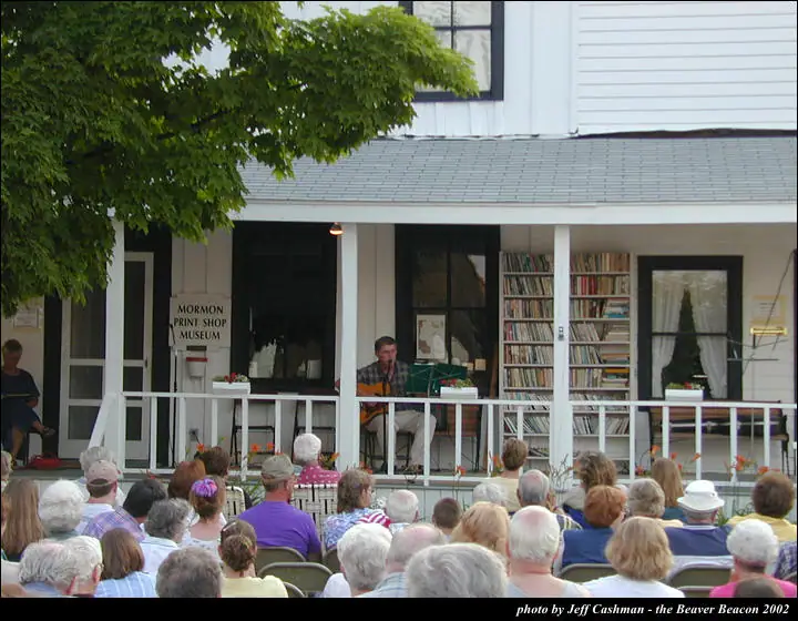 A guitarist performs on a porch before a seated audience.