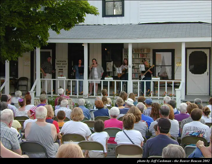 Musicians perform on a covered porch before a large seated audience.