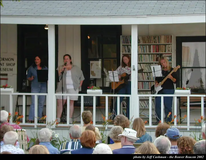 Four women perform with microphones and guitars on a porch before a seated audience.