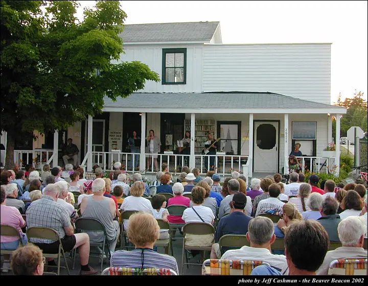 A band performs on a covered porch before a large seated audience.
