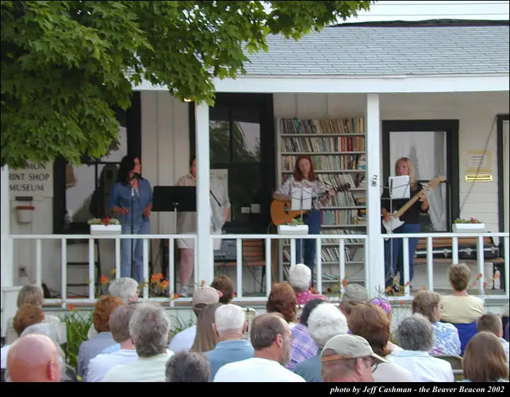 Musicians perform on a porch before a seated audience, with bookshelves and a museum sign behind them.