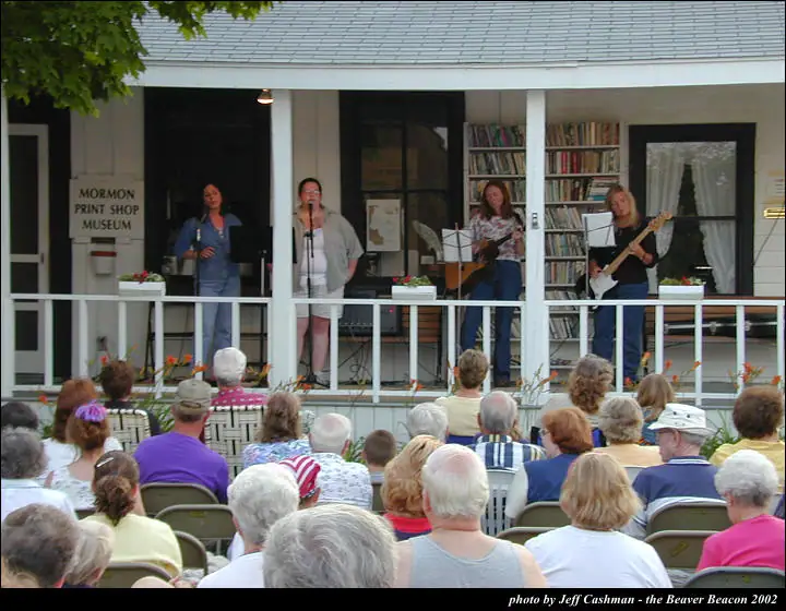 Four women perform on a porch before a seated audience, with guitars, microphones, and a museum sign visible.