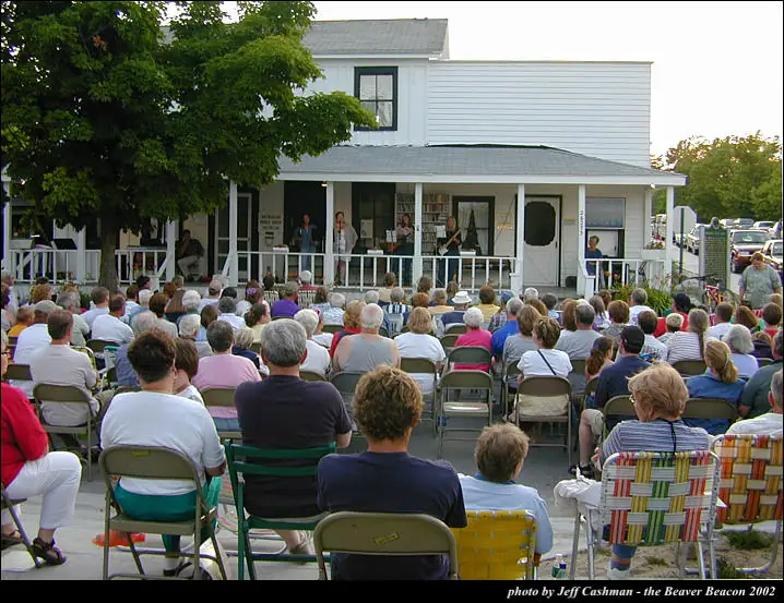 A large seated audience watches musicians performing on the porch of a white building.