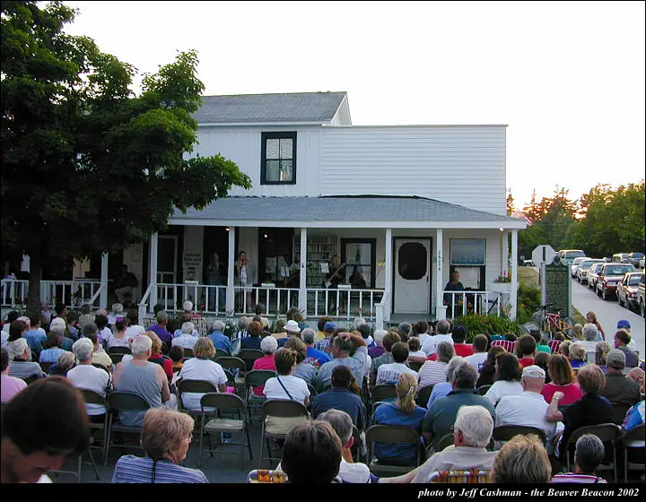 A large seated crowd watches musicians performing on the porch of a white building.