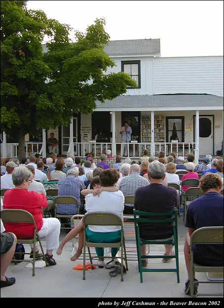 A seated crowd faces a speaker standing on the porch of a white building.