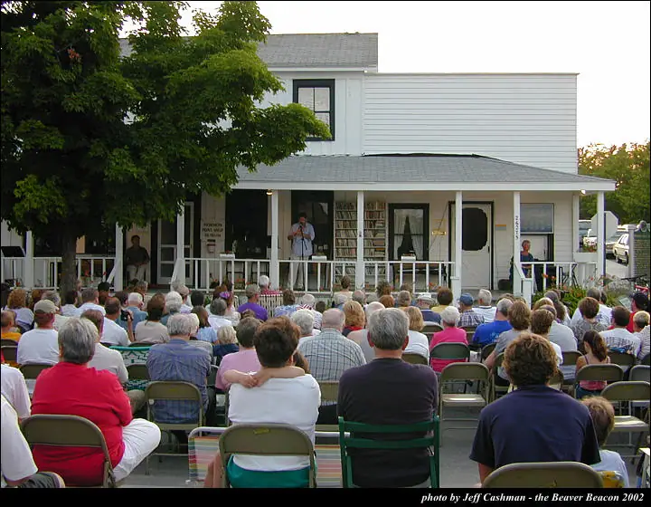 An outdoor audience watches a person speaking at a microphone from a covered porch.