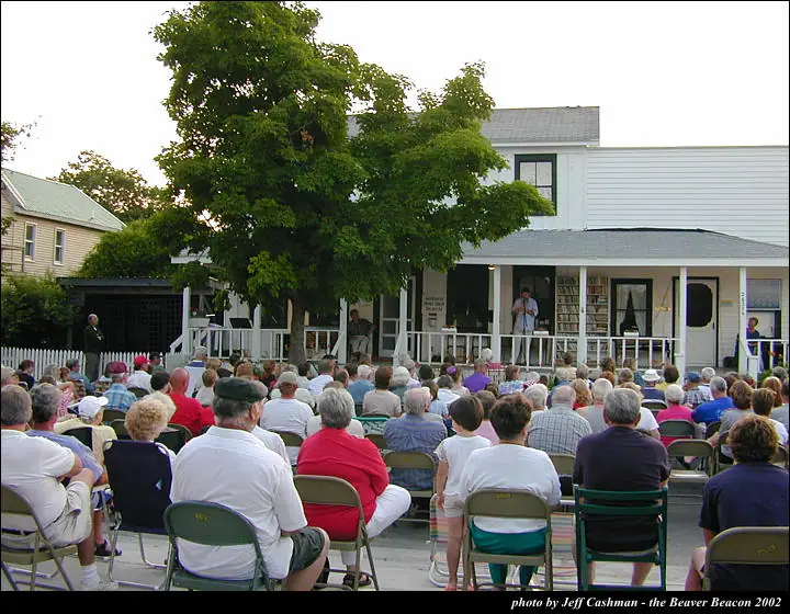 A large audience sits in folding chairs facing a person on a covered porch beside a leafy tree.