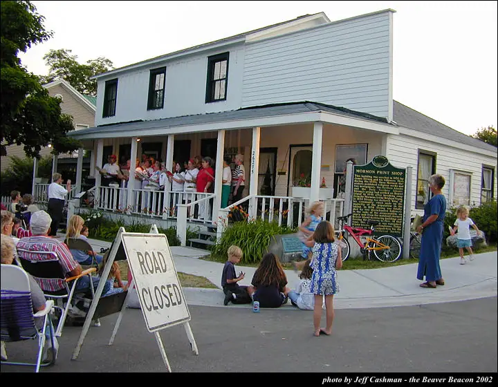 A group performs on a porch while people watch from the sidewalk beside a building marked as a print shop.