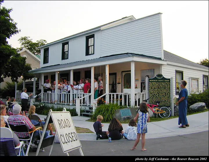 A group stands on a porch while spectators gather outside a white building beside a road-closed sign.