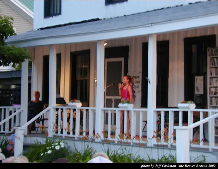 A girl sings into a microphone on a flower-decorated porch while people watch.