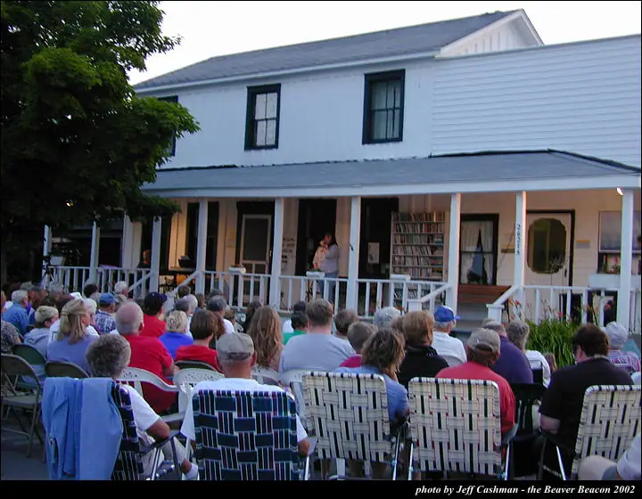 A crowd seated in lawn chairs faces a person on the porch of a white two-story house.