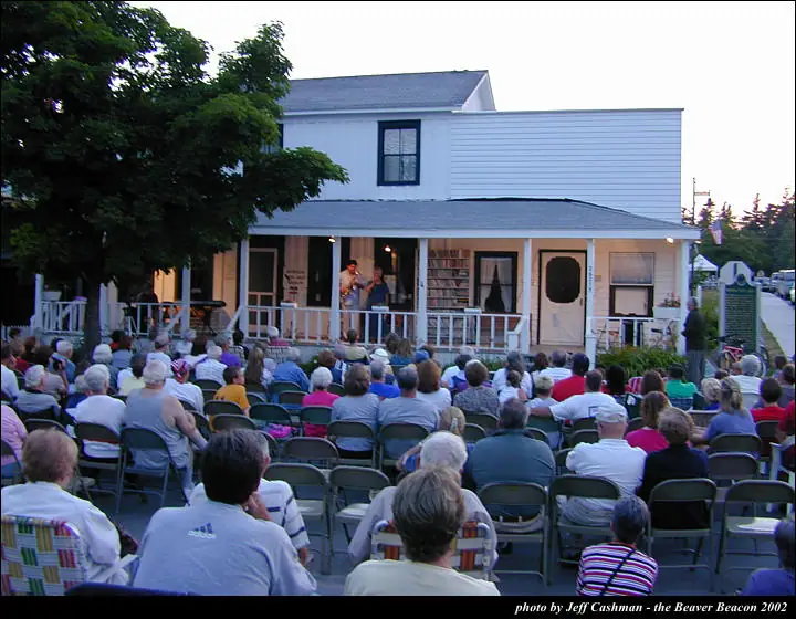 A crowd watches two musicians perform from the porch of a white two-story building.