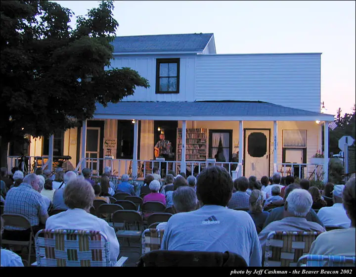 A guitarist performs on a house porch before a large seated outdoor audience.