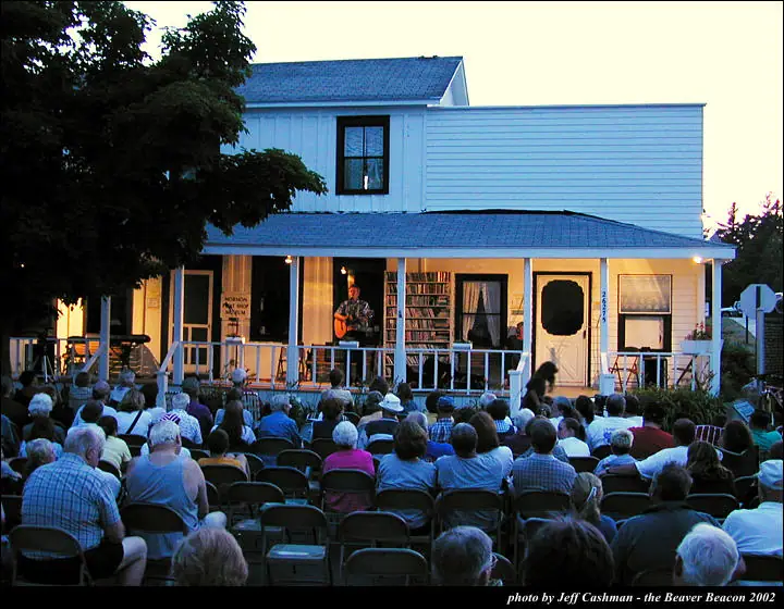 A guitarist performs on a lit porch before a large seated audience.