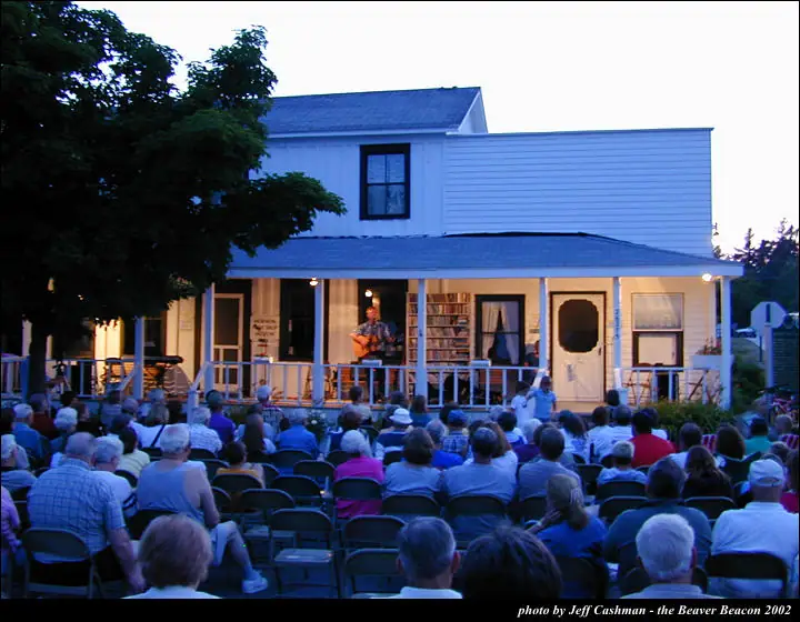 A guitarist performs on a house porch while a large seated audience watches at dusk.