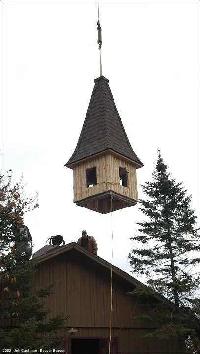 A wooden bell tower hangs above a building roof while a person stands below amid evergreen trees.