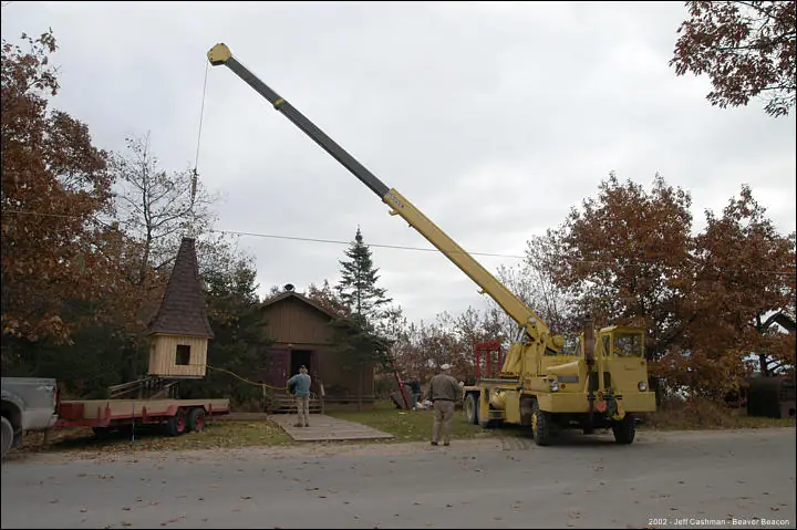 A crane lifts a small wooden steeple beside a trailer and cabin.