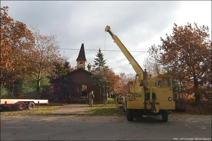 A yellow crane stands beside a small wooden church with a pointed steeple and several people nearby.