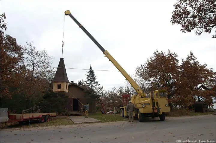 A crane lifts a wooden church steeple beside a small church as workers stand nearby.