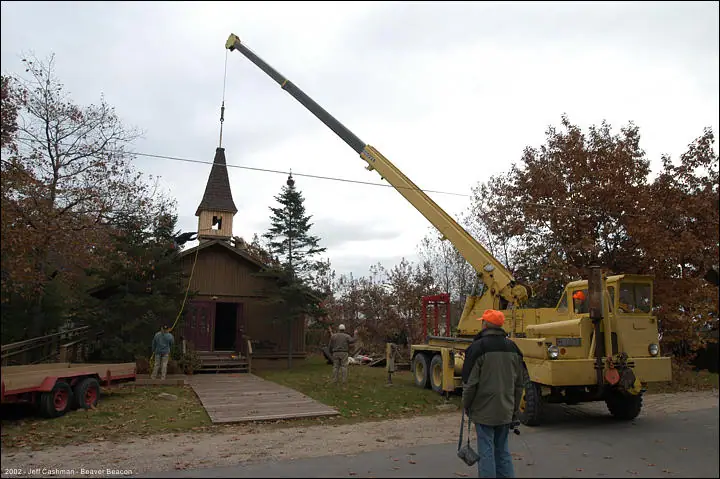 A crane lifts a steeple onto a small wooden church while people watch.