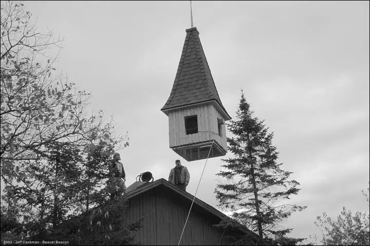 Black-and-white photograph of a steeple suspended above a wooden building while two people work below.