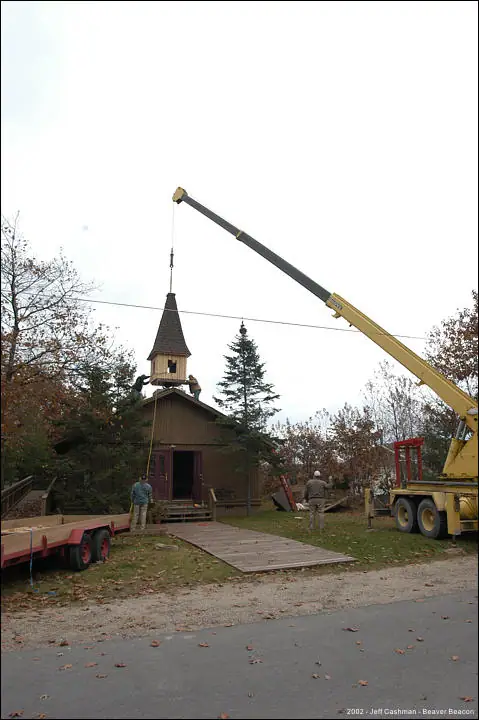 A crane lifts a pointed steeple above a small wooden church as people watch.