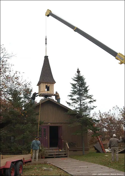 A crane lifts a wooden steeple onto a small wooden church.
