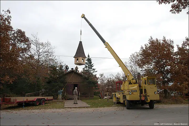 A crane lifts a steeple above a small wooden church while workers stand nearby.