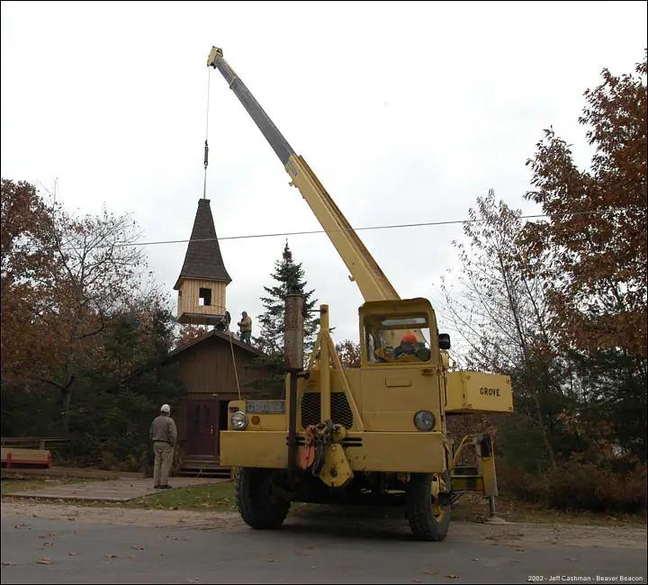 A yellow crane lifts a pointed wooden steeple above a small church as workers guide it into position.