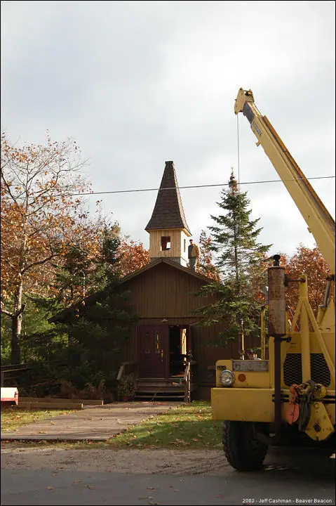 A yellow crane stands beside a small wooden church with a steeple and surrounding trees.