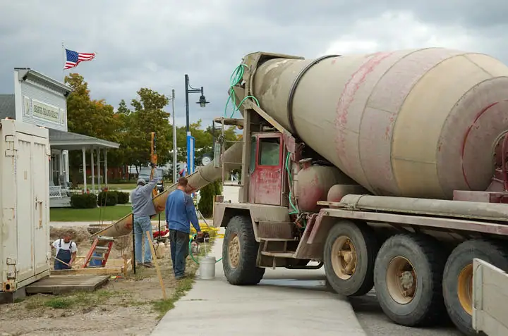 Workers pour concrete from a mixer truck into a foundation trench beside a building.