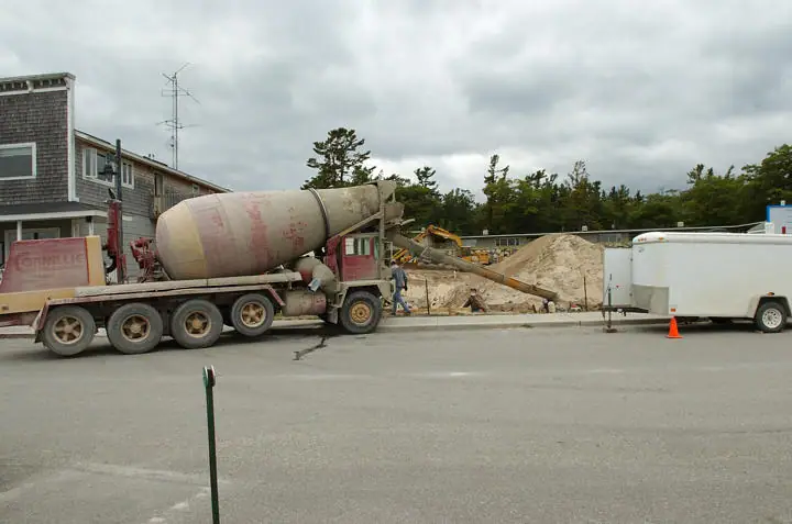 A concrete mixer truck and workers pour concrete at a construction site beside a white utility trailer.