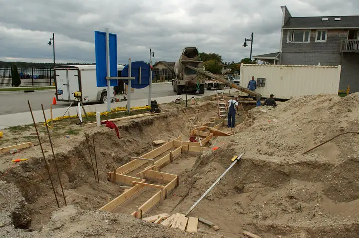 Workers prepare wooden forms in a foundation excavation beside a concrete mixer truck.