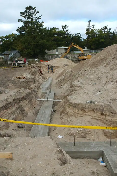Construction site with concrete footings, exposed rebar, an excavator, sand piles, and yellow caution tape.