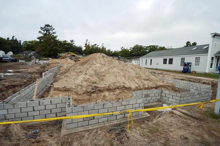 Concrete block foundation walls surround a large sand pile at a construction site, with an excavator and buildings behind.