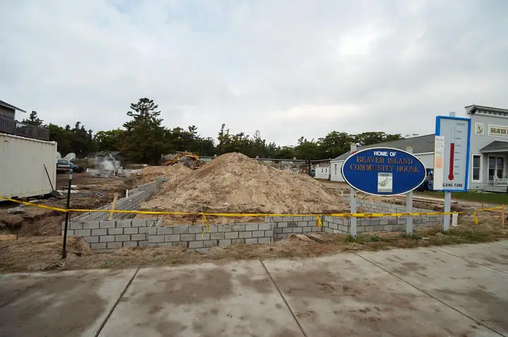 Construction site with a low concrete-block foundation, sand pile, caution tape, and a sign for a community house.
